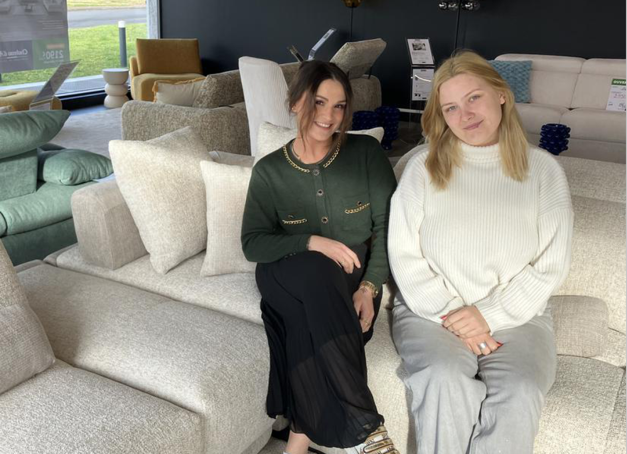 Deux personnes souriantes sont assises sur un canap beige dans un salon moderne avec plusieurs fauteuils et coussins autour.  Description g n r e par IA