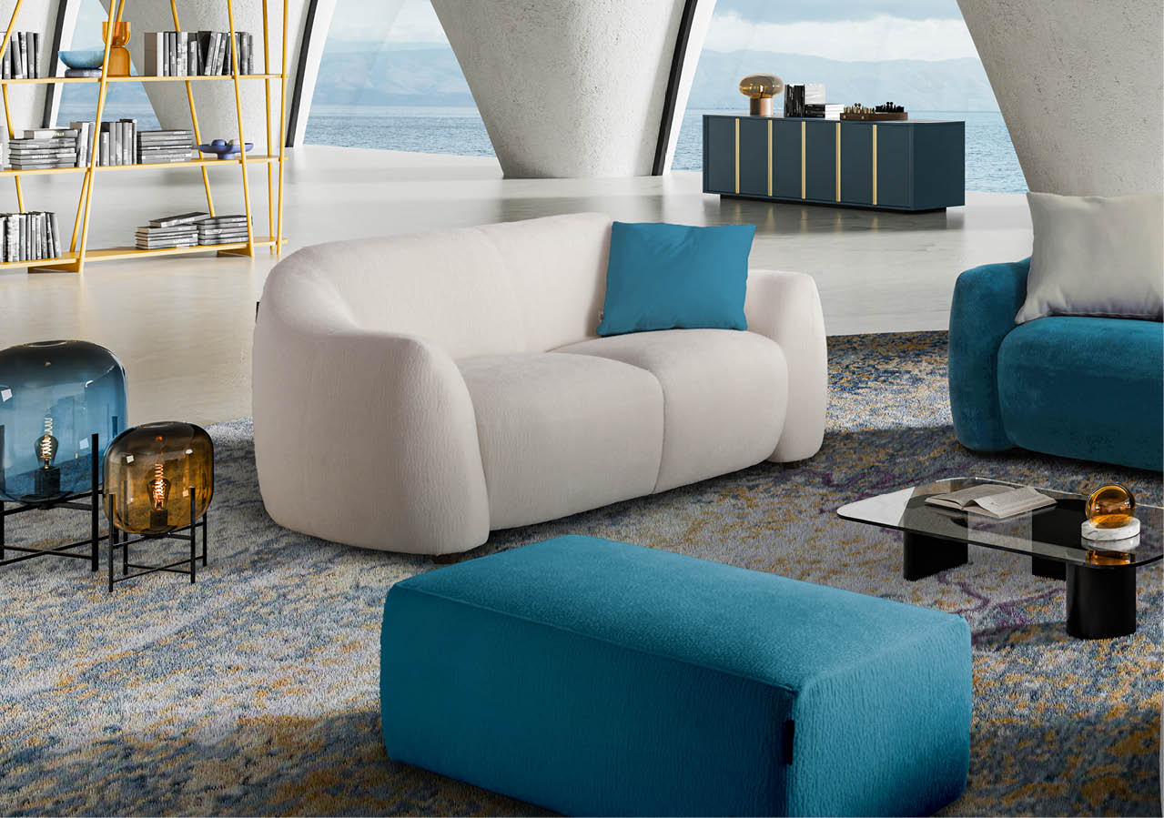 Un salon moderne avec un canap beige, un pouf bleu, une table basse en verre, des lampes d coratives et une grande fen tre offrant une vue sur la mer.  Description g n r e par IA
