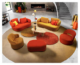 Un salon moderne avec deux canap s en cuir marron, un fauteuil assorti, une table basse noire sur un tapis bleu et deux poufs avec un livre ouvert, le tout sur un sol en bois clair pr s d'une grande fen tre. Description g n r e par IA