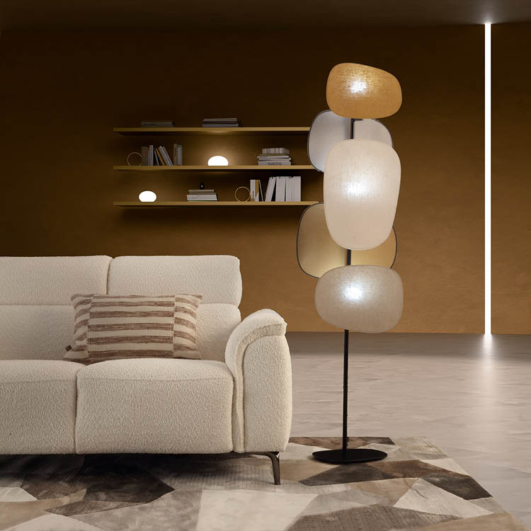 Un salon moderne avec un canap beige, un coussin ray , une lampe sur pied design compos e de plusieurs abat-jours ovales et des  tag res murales avec des livres et des objets d coratifs sur un mur marron.  Description g n r e par IA