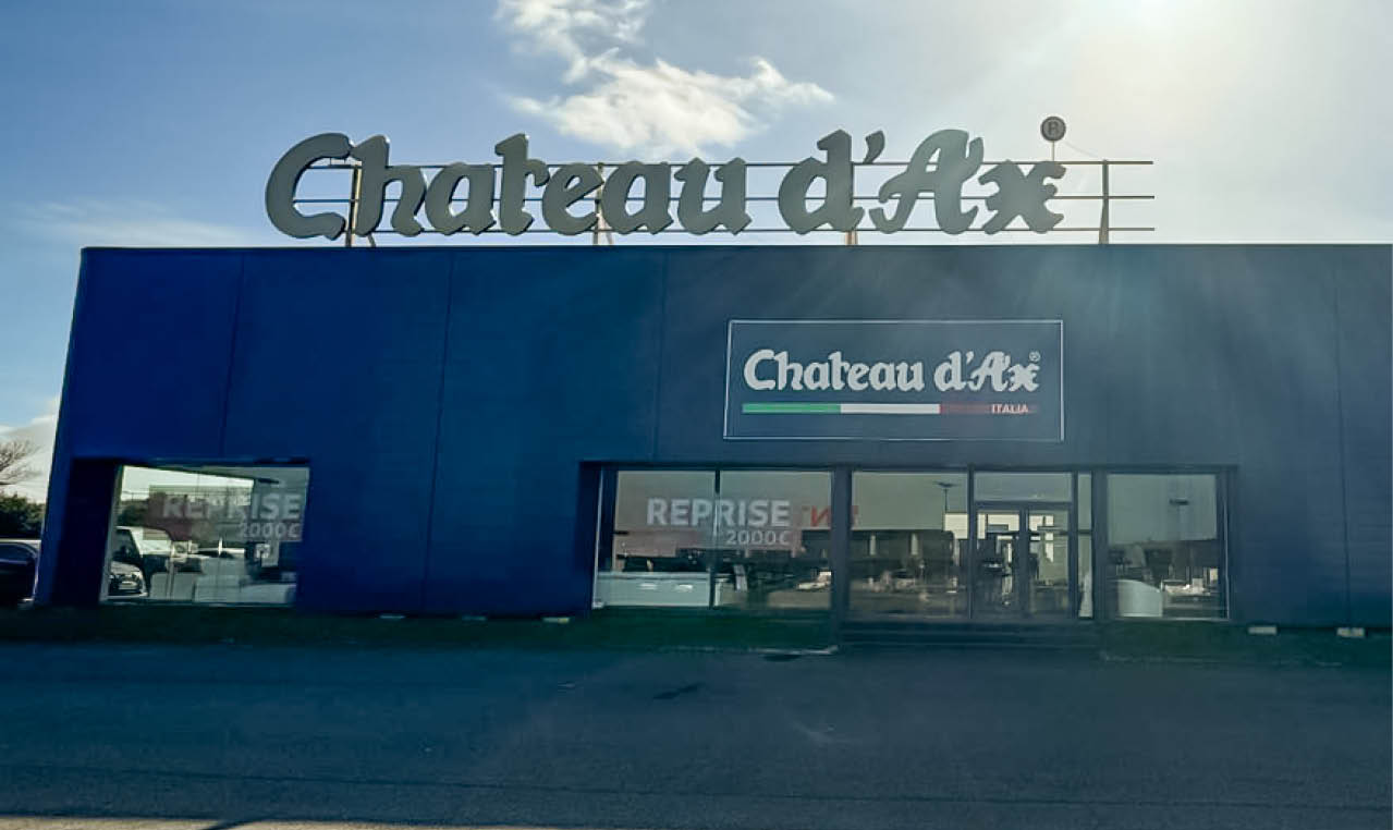 B timent commercial bleu avec une grande enseigne “Chateau d'Ax" sur le toit et une vitrine avec le m me nom affich . Description g n r e par IA