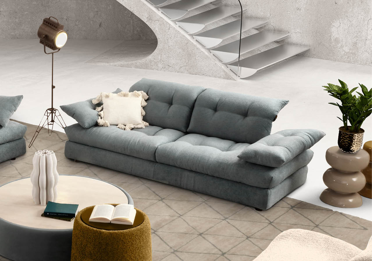 Un salon moderne avec un canap gris clair orn  de coussins, une lampe sur pied vintage, une table basse ronde avec des livres et un tabouret jaune moutarde, le tout sur un tapis   motifs g om triques devant un escalier en colima on.  Description g n r e par IA