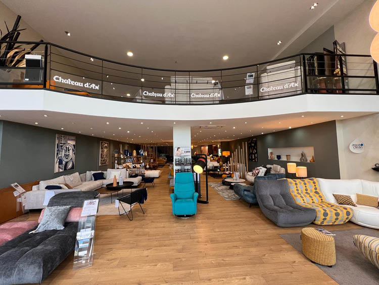 Un grand showroom lumineux pr sente plusieurs canap s modernes et color s dispos s sur un sol en bois avec une mezzanine et des enseignes Ch teau d'Ax. Description g n r e par IA