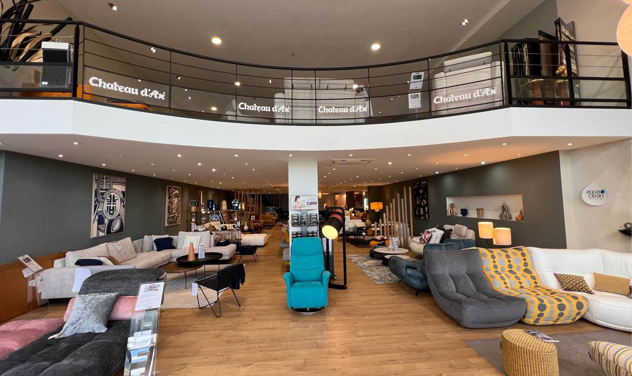 Un grand showroom moderne pr sente plusieurs canap s et fauteuils color s dispos s sur un sol en bois avec un balcon et des enseignes lumineuses Ch teau d'Ax en arri re-plan. Description g n r e par IA