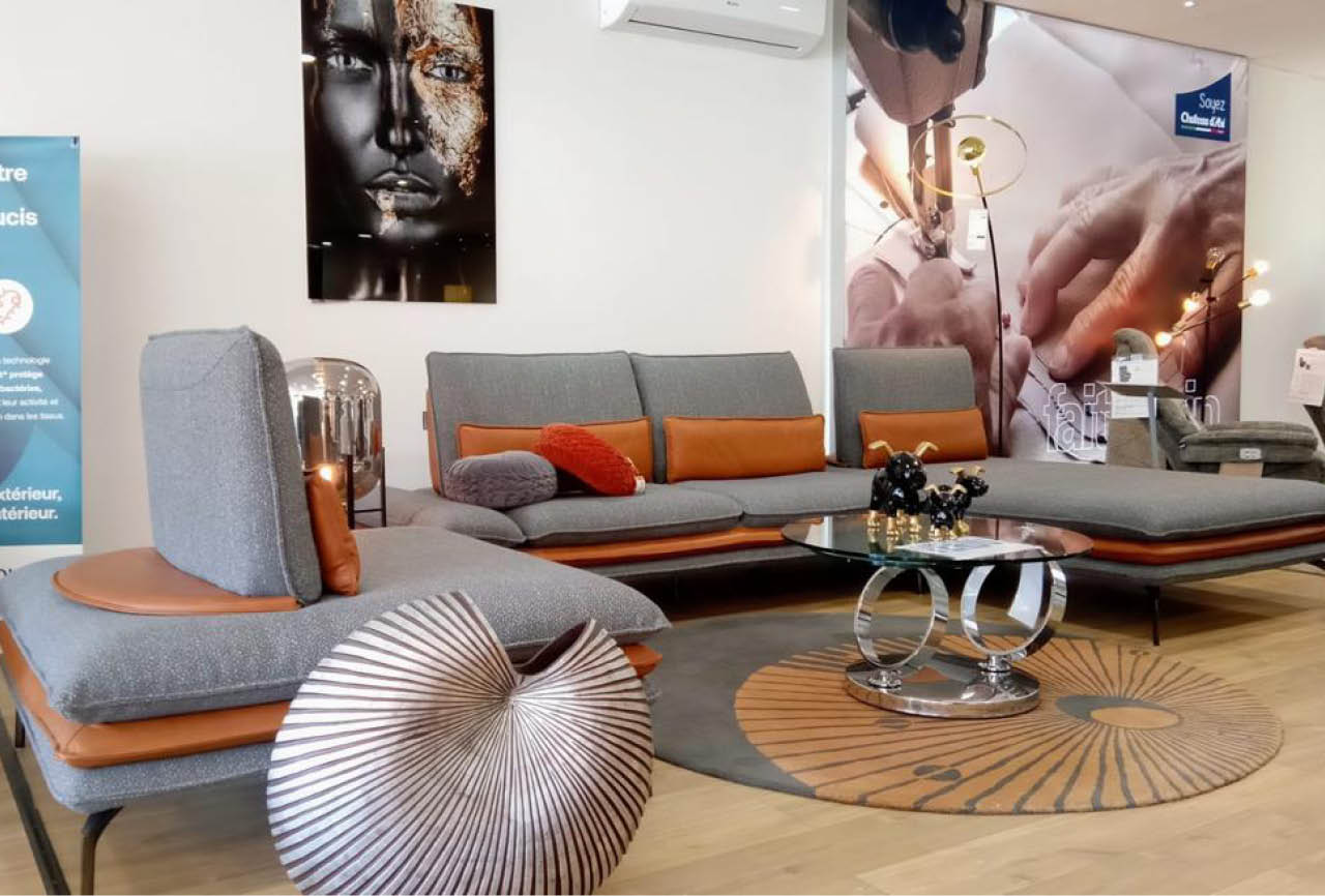 Un salon moderne avec un canap modulable gris et orange, une table basse en verre avec des d corations, un tapis rond et une grande affiche murale montrant des mains travaillant sur un objet.  Description g n r e par IA