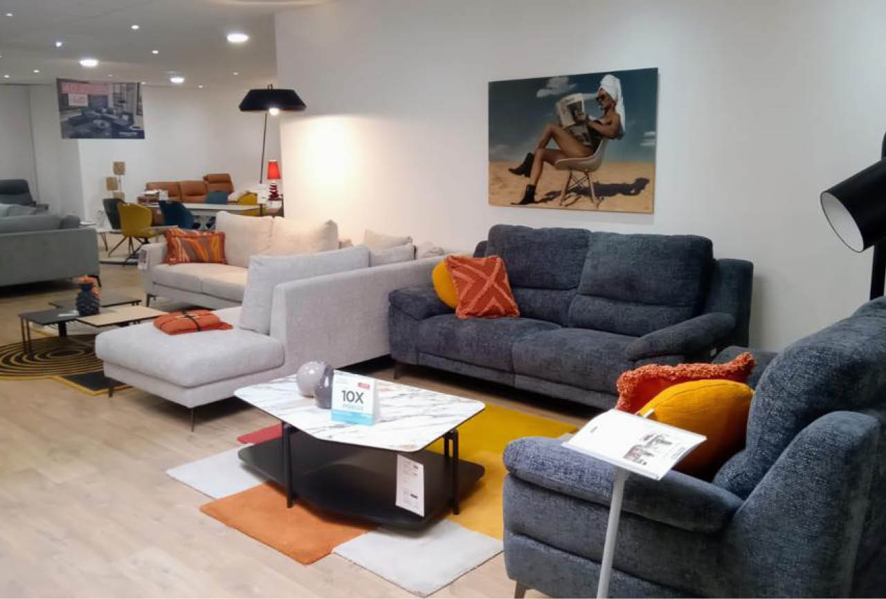 Un salon moderne avec plusieurs canap s gris et bleu, des coussins color s, une table basse avec un panneau d'information, et un tableau repr sentant une personne assise sur une chaise au bord de la mer. Description g n r e par IA