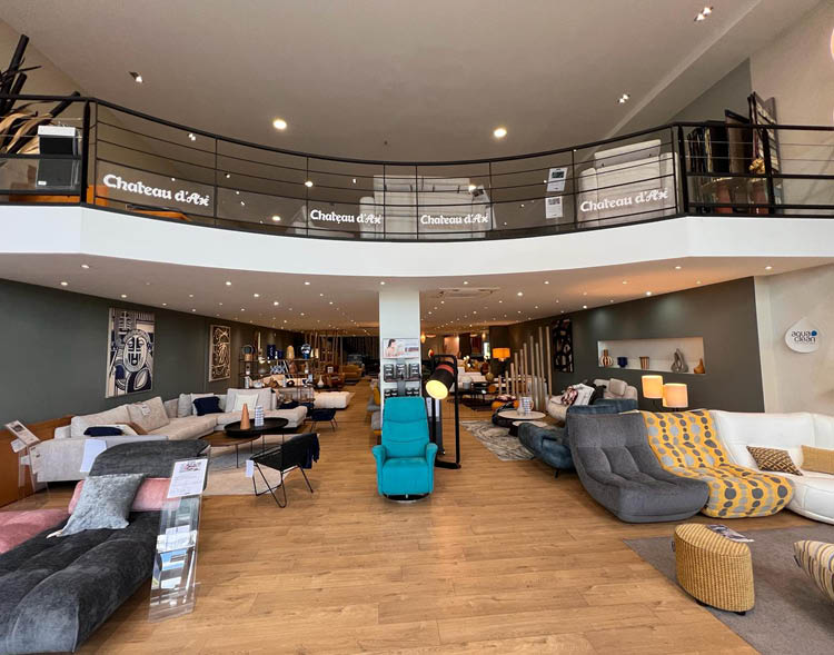 Un grand showroom moderne pr sente plusieurs canap s et fauteuils color s dispos s sur un sol en bois clair avec un balcon en mezzanine et un clairage chaleureux.  Description g n r e par IA